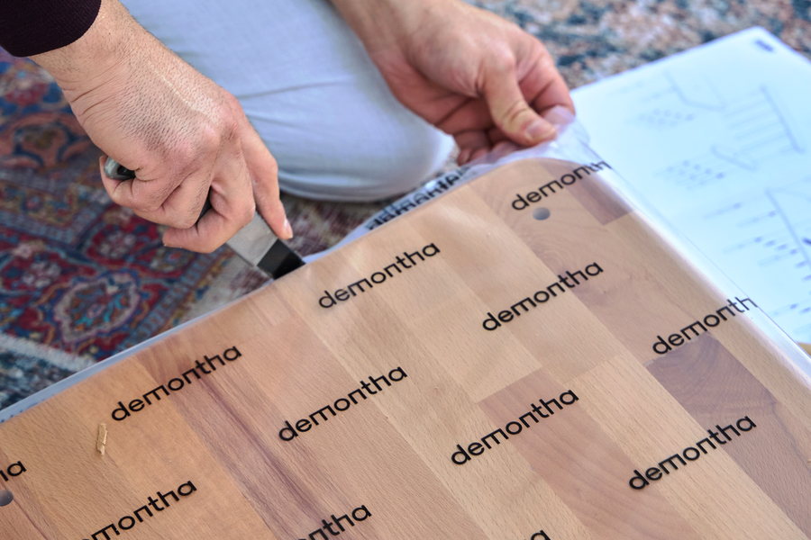 A person kneeling on a rug opens a thick solid beech panel wrapped in demontha-printed protective film; the lamella structure shows through the film