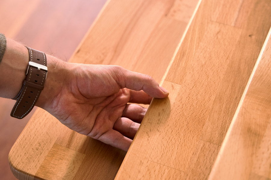 A hand resting on the front edge of a solid beech bench; the rounded edge profile and the lamellas joined side by side are seen up close